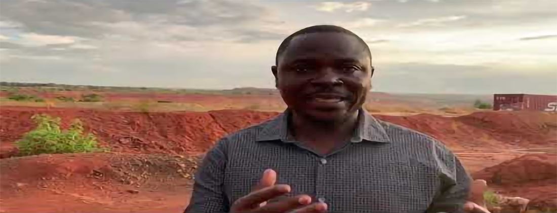 A man speaks to camera in front of a red-earth open-pit mining landscape in the DRC