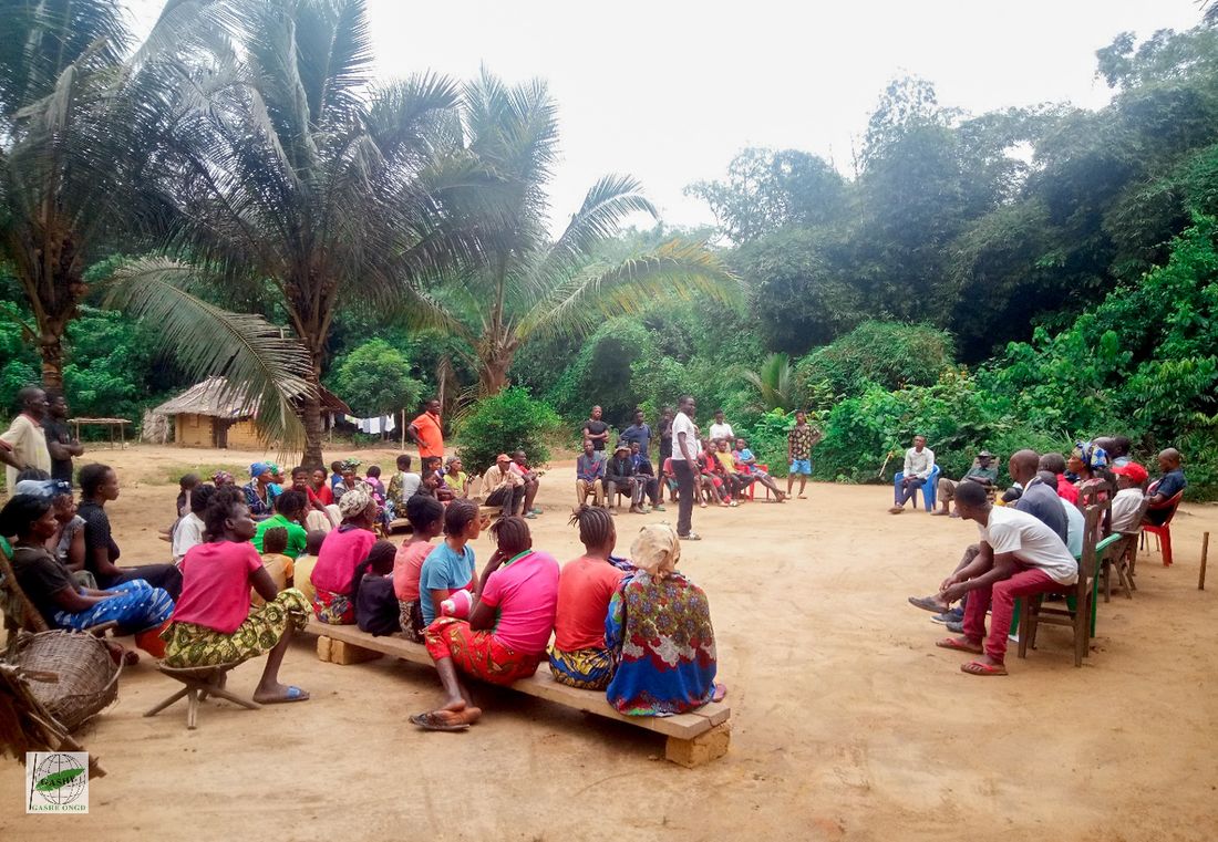 A facilitator addresses a community gathering in a forest village clearing in Équateur
