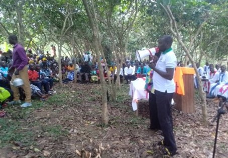 A speaker with a megaphone addresses a community gathering under the trees in Mahagi